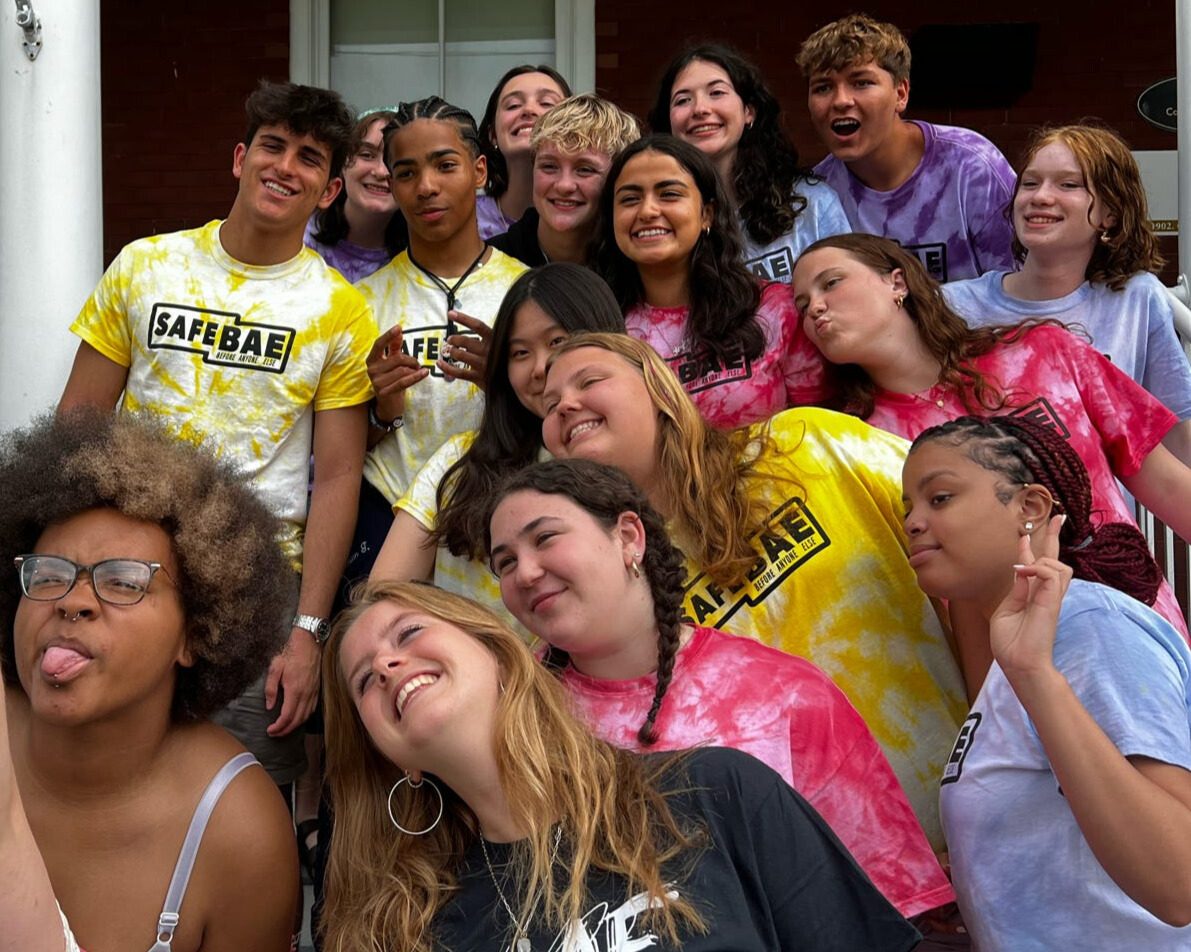 A group of SafeBAE students in tie-dye SafeBAE shirts, smiling and making faces on a front porch